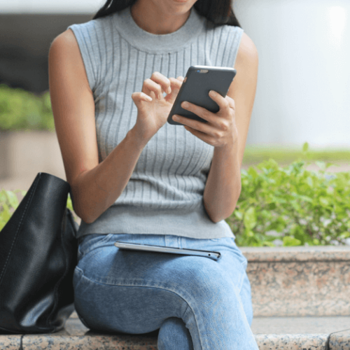 Imago of woman in a grey turtleneck and jeans scrolling on her phone with a tablet in her lap