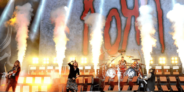 Image of Korn performing on stage surrounded by smoke columns and lights with a gray sign with 'Korn' in big, red letters on it