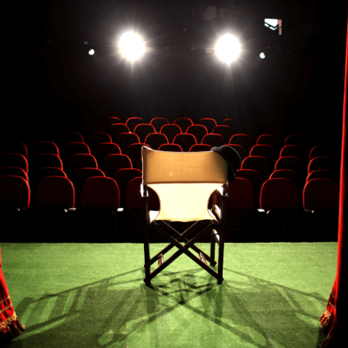 Image of director's chair with a bowler hat in the spotlight on stage facing red theater seats
