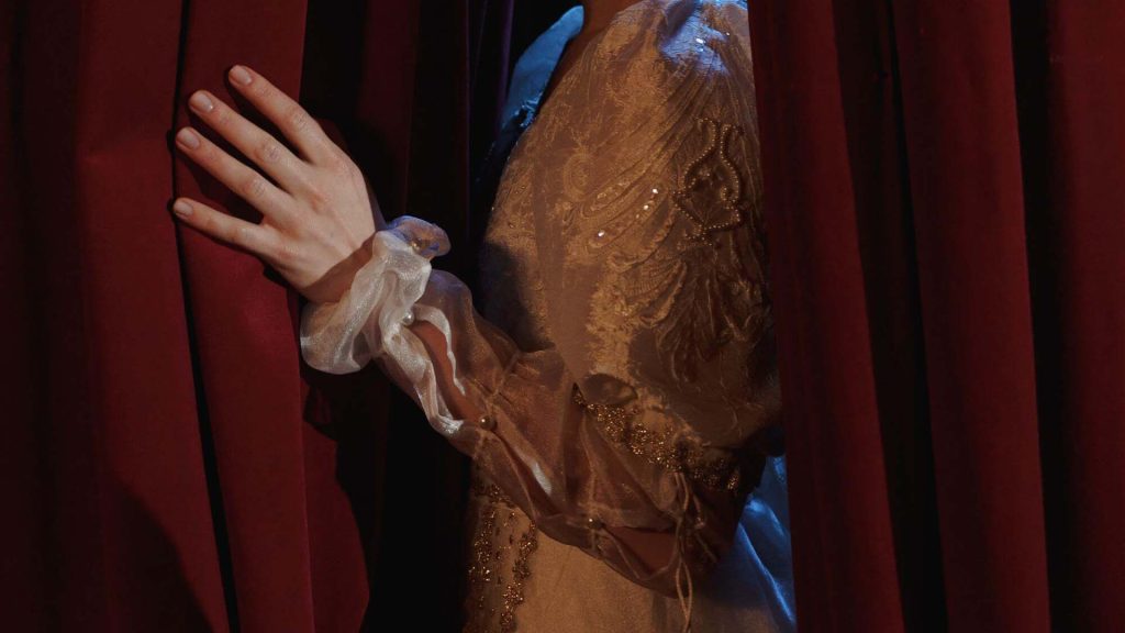 An arm dressed in older styled closed pulls back a theatre curtain.