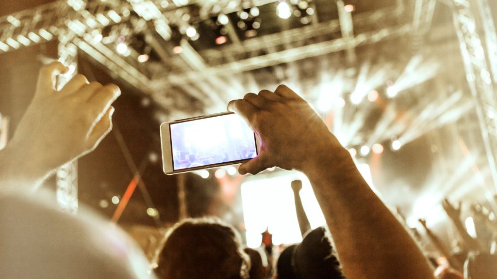 Photo of an arm holding up a phone in front of a stage with bright lights.