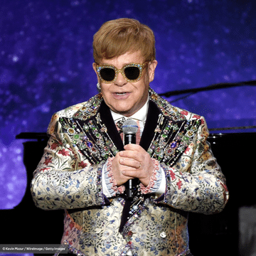 Elton John wearing sunglasses and stnading in front of a purple background