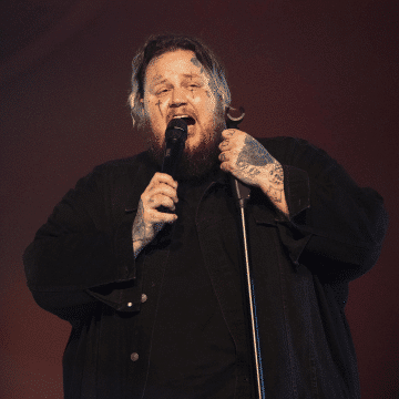 Jelly Roll wearing a black denime jacket and shirt singing into a microphone