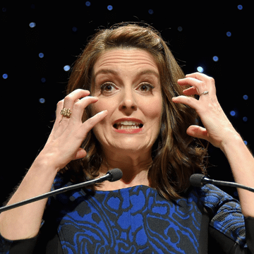 Photo of Tina Fey in front of a black backround. There is a microphone in front of her and she has her hands up near her face.