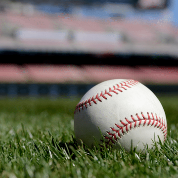 Photo of a white baseball with red stitching sitting on a green grass field. There are red and silver bleachers blurred in the background.
