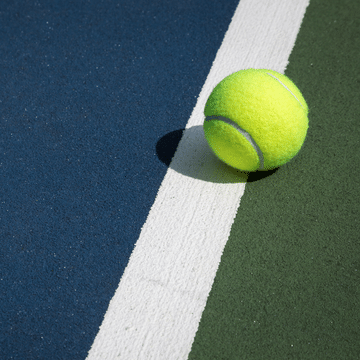 Photo of yellow tennis balling sitting on a white line of a hard two-tone blue and green tennis court.