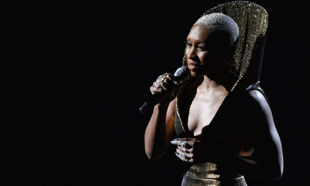Cynthia Erivo Singing under a spotlight wearing a gold dress with a high collar