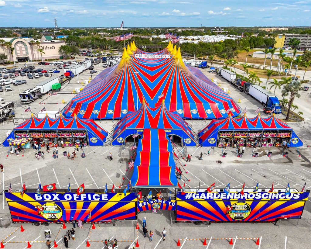 Photo of the Garden Bros Nuclear Circus tent from above. Tent has red and club stripes with gold caps.