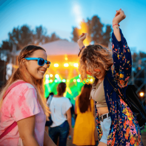 Two young women wearing colored sunglasses dance and smile at a music festival.