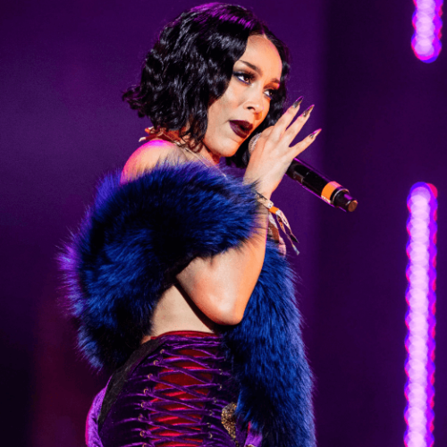 Image of Doja Cat singing while wearing a purple furred shawl and a dark pink mini skirt with black laced sides against a purple stage background