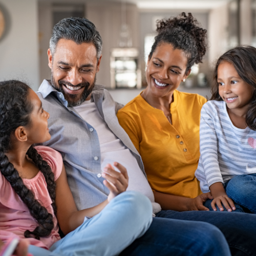 Photo of a family sitting on a couch and smiling while listening to a young girl talk.