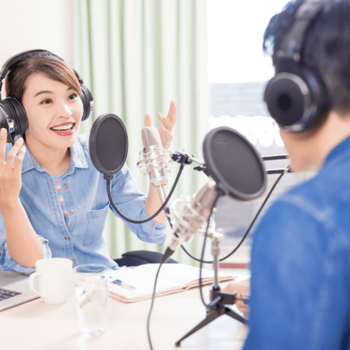 A woman wearing headphones and a blue shirt sitting at a table with a man wearing headphones and a jean jacket speaking into podcast microphones