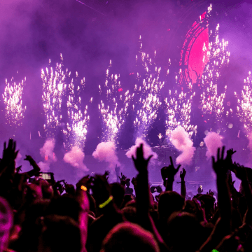 A crowd waves their hands in their air as fireworks go off on stage in front of them.
