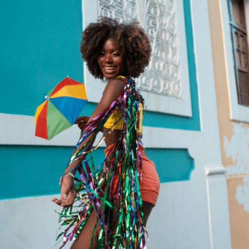 Image of woman standing in front a blue wall wearing a yellow top and shorts holding a rainbow umbrella with colorful tinsel draped down her arm and shoulders