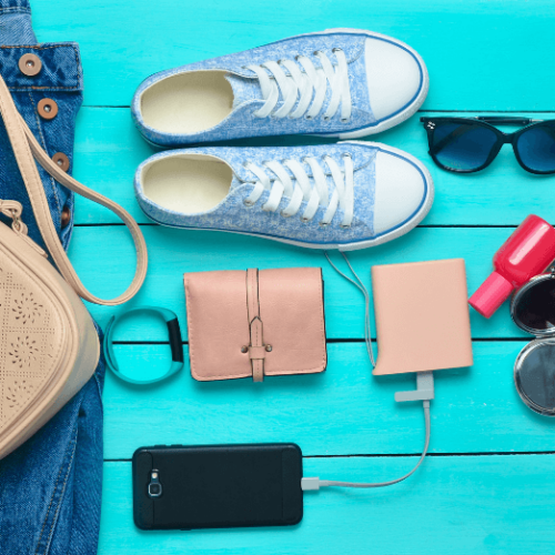 Image of various items against a bright blue background from above including blue tennis shoes, sunglasses, tan wallet, black smartphone connected to a power bank, smart watch and pink bottle of nail polish
