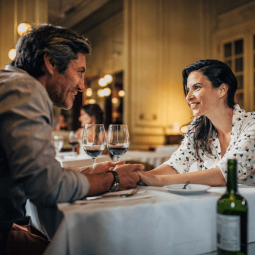 Image of a couple sitting a dinner table holding hands with two glasses of wine