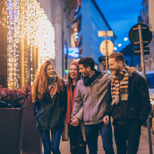 Image of a group of four people walking together on the street past a storefront with twinkle lights and a planter with pink flowers