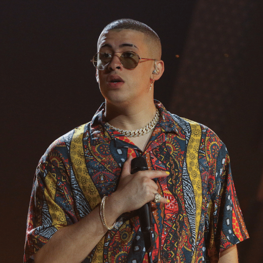 Photo of Bad Bunny holding a microphone with one hand. He has a multi-colored shirt and sunglasses on.