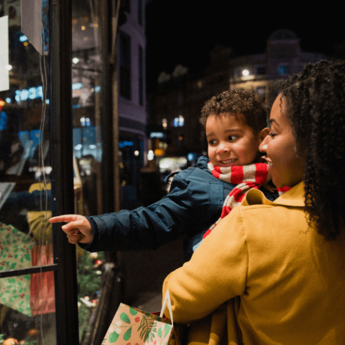 Photo of a woman holding a young child. They are looking through a store window and the child is pointing at it.