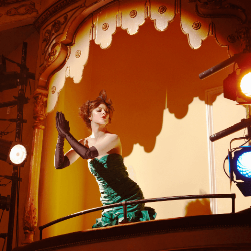 Image of woman in a strapless green dress and black opera gloves standing on a theatre balcony