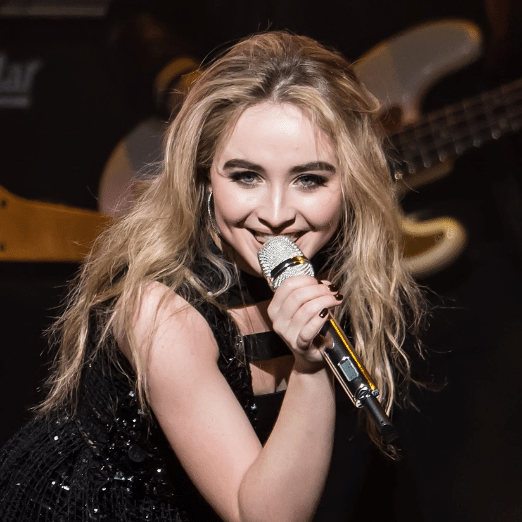 Photo of Sabrina Carpenter holding a microphone and singing into it. She is facing directly into the camera.